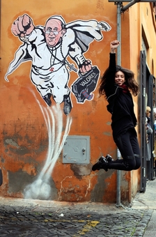 دختری در مقابل نقاشی دیواری از پاپ به تقلید از سوپرمن در حال پریدن است و دوستانش از او عکس می گیرند. در این نقاشی دیواری، پاپ فرانچسکو شبیه به سوپرمن ترسیم شده که در حال پریدن به آسمان است/euronews

